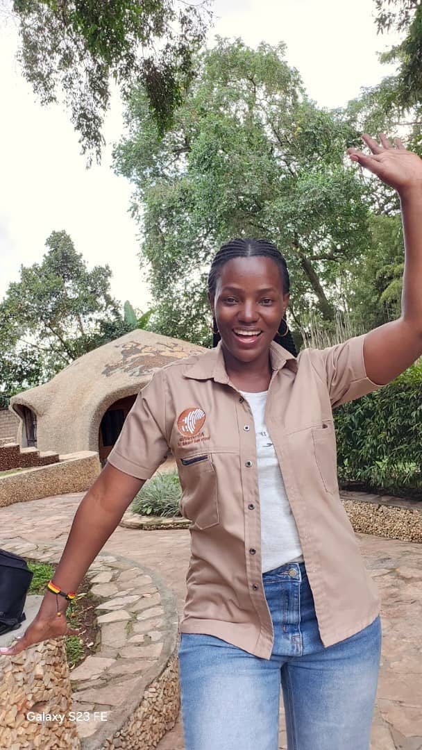 Smiling woman in tan uniform shirt and jeans standing outdoors with arm raised, trees and thatched structure visible in background
